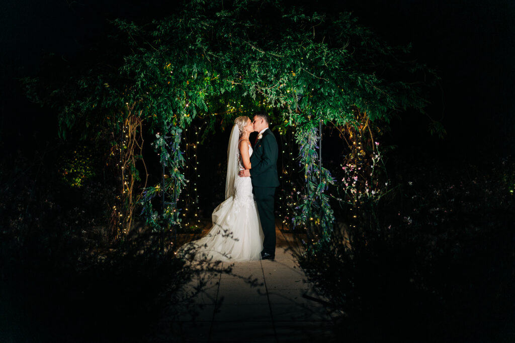 A bride in a white gown and a groom in a black suit kiss under a beautifully lit pergola adorned with greenery and twinkling lights at night.