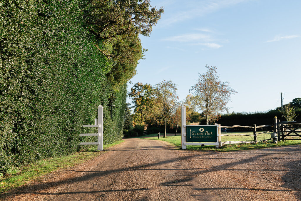 A gravel road leads into a park, flanked by tall green hedges and trees. A sign on the right reads "Gaynor Park, Recreational Grounds." The sky is clear, and sunlight illuminates the scene, creating a peaceful, rural atmosphere.