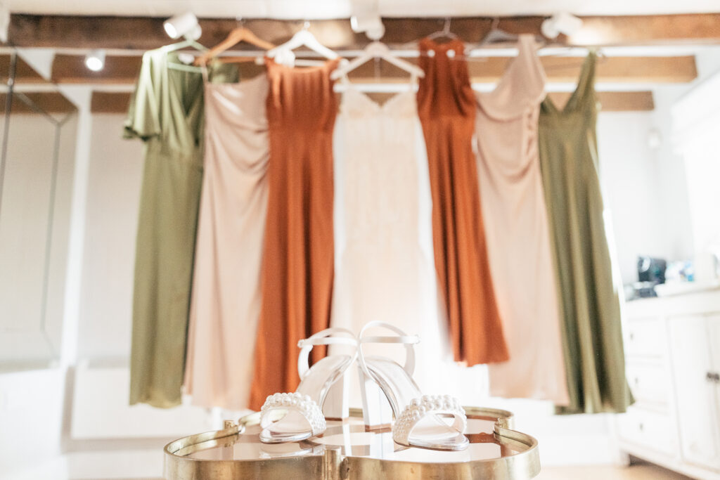 A pair of elegant white heels with pearl details is placed on a round table in the foreground. In the background, five dresses in earthy tones, including green, orange, and beige, are hung on wooden hangers, showcasing a bridal party setup.