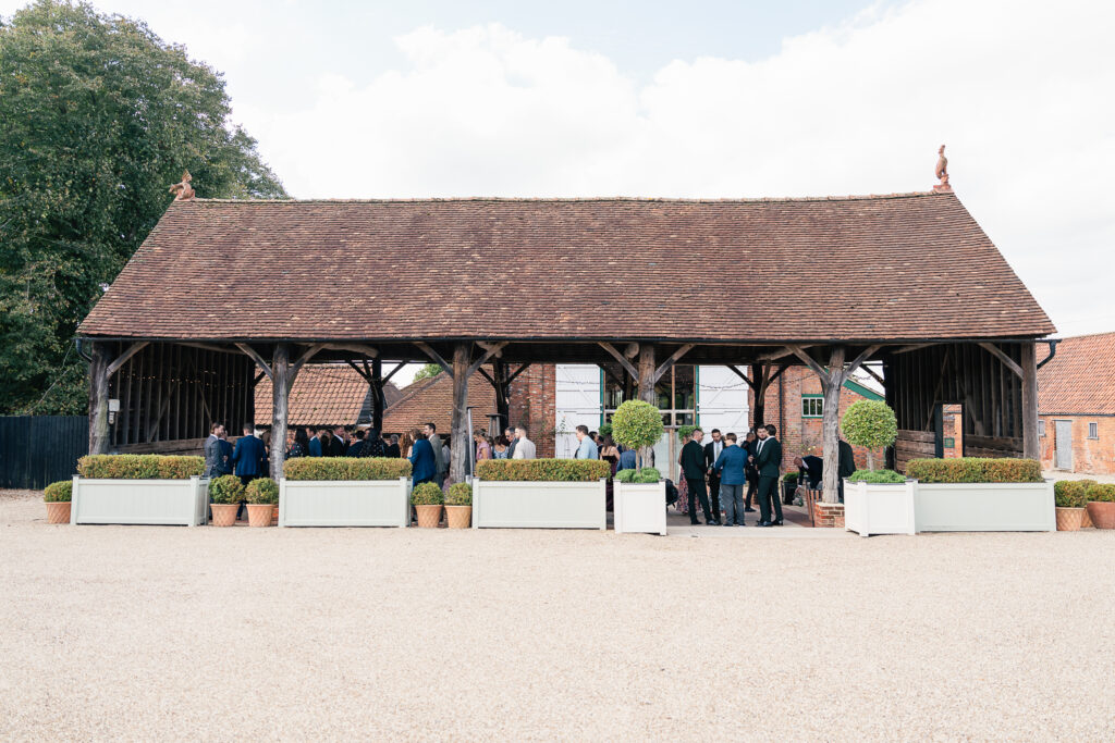 Outdoor gathering under a wooden barn structure with a red-tiled roof. People are mingling beside large planters with greenery on a gravel surface. Trees are visible in the background.