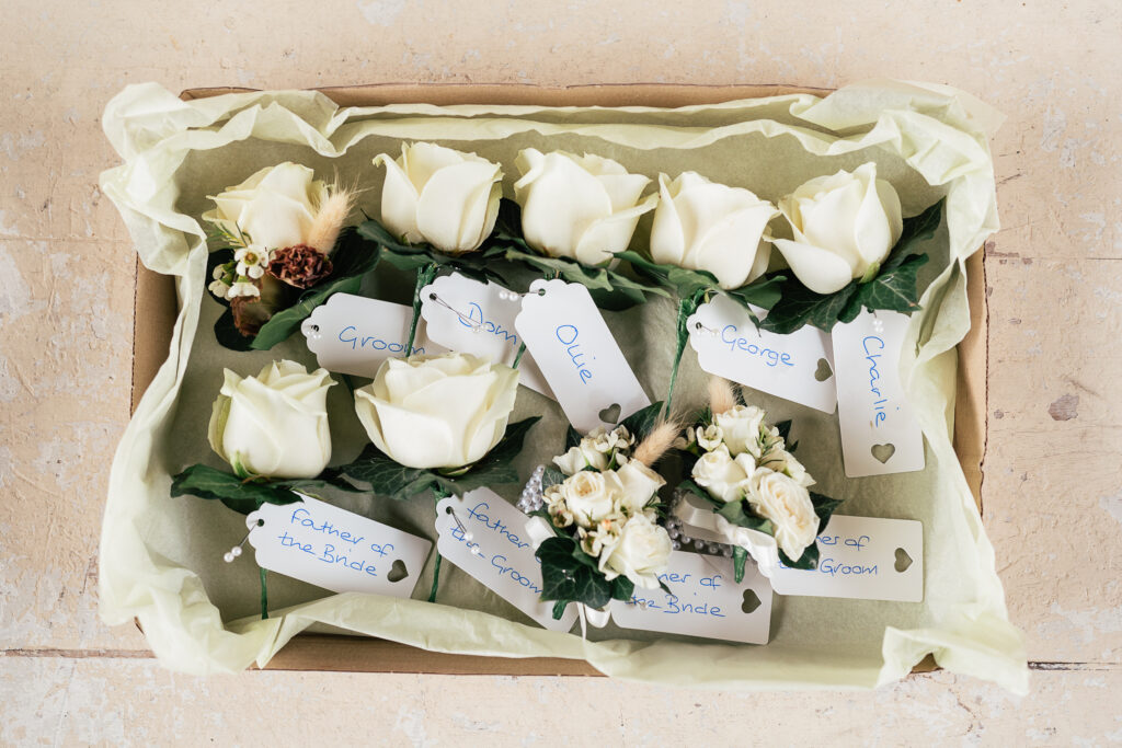 A box filled with white rose boutonnieres, each labeled with names such as "Groom," "Dad," "George," and roles like "Father of the Bride," arranged on light green tissue paper.