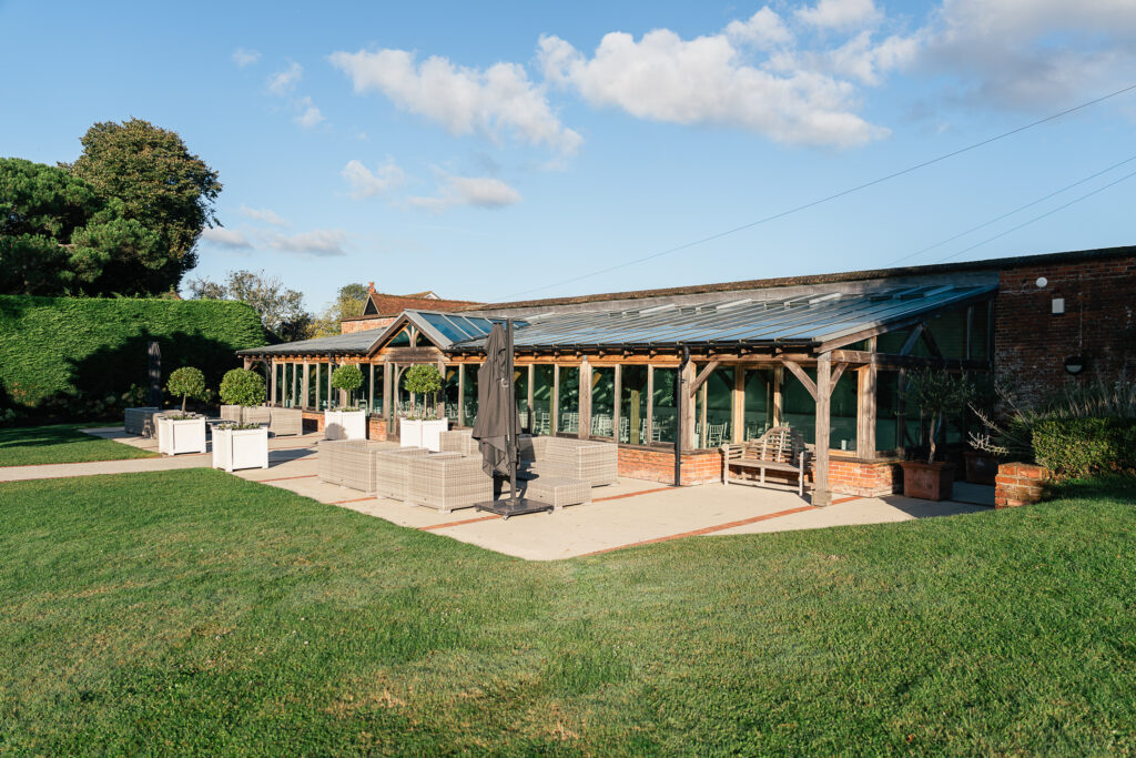 A glass and wood outdoor pavilion surrounded by neatly trimmed grass and shrubs. The structure has seating areas with contemporary furniture, including chairs and tables. A partly cloudy sky is visible above.