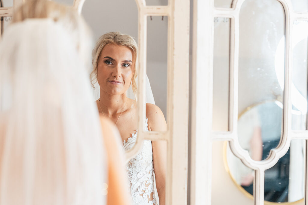 A bride in a white lace wedding dress and veil looks at her reflection in a decorative mirror. The setting appears calm, with soft lighting enhancing the gentle expression on her face.