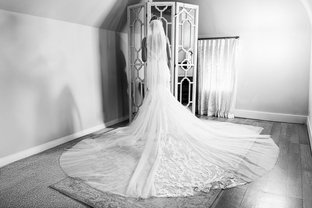 A black and white image of a bride standing in front of a decorative mirror, wearing a lacy wedding gown with a long train flowing behind her. The room has wooden flooring and soft lighting, with a curtain-covered window to the side.