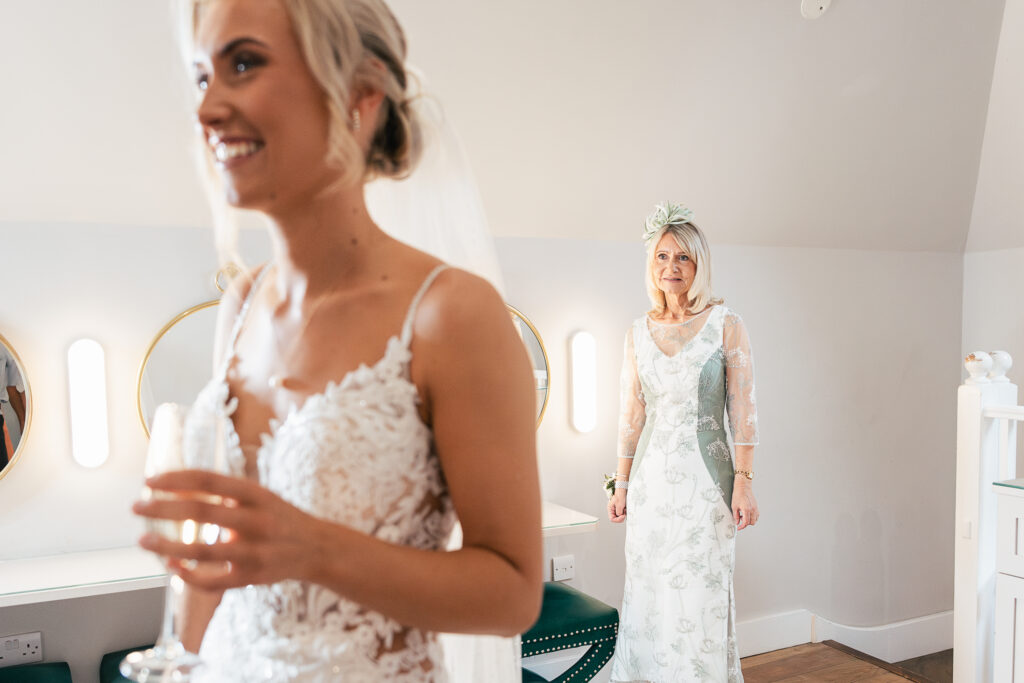 A bride in a lace wedding dress smiles in the foreground, holding a glass. In the background, another woman in a floral dress and hat looks fondly at the bride. The room is softly lit with round mirrors and modern decor.