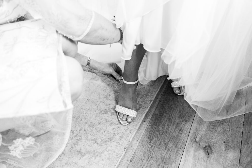 Black and white image of a person fastening a strap on a bride's high-heeled shoe, with the lower part of her dress visible. The setting appears to be indoors on a wooden floor and a rug.