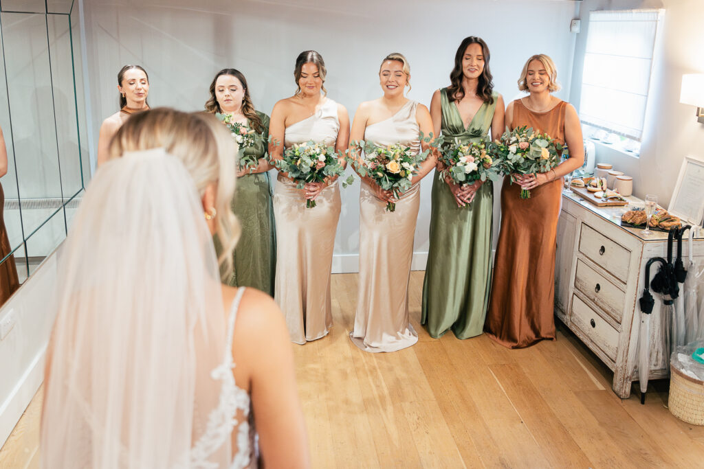 A bride stands with her back to the camera, facing six bridesmaids. The bridesmaids, dressed in various shades of green and beige, hold bouquets of flowers. They are smiling in a room with hardwood floors and soft lighting.