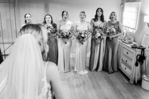 A bride, seen from behind, stands facing a group of six bridesmaids indoors. The bridesmaids, dressed in elegant gowns and holding bouquets, display joyful and surprised expressions. The room features wooden floors and soft lighting.