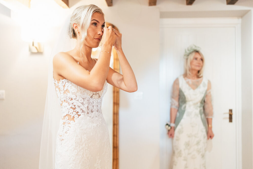 A bride in a lace wedding dress adjusts her earring while standing in a softly lit room. In the background, a woman in a floral dress and matching fascinator watches.