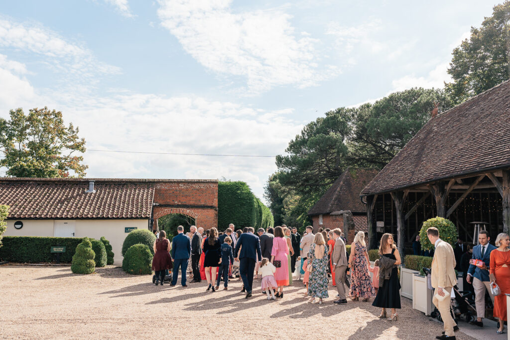 Guests in formal attire gather outside a rustic brick barn under a partly cloudy sky, surrounded by well-maintained greenery.