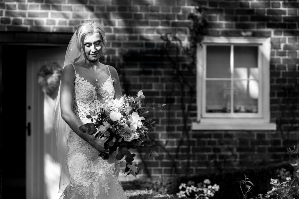 A bride in a lace wedding dress stands outside, holding a bouquet of flowers. She is in front of a brick building, with light and shadow patterns across her. A heart-shaped wreath hangs on the door behind her.