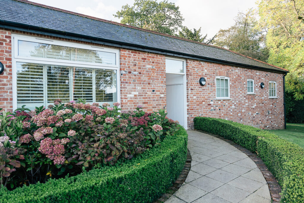 A charming brick house with large windows and a slate roof, surrounded by green hedges and blooming hydrangeas. A curved stone path leads to the entrance, flanked by manicured greenery and trees in the background.