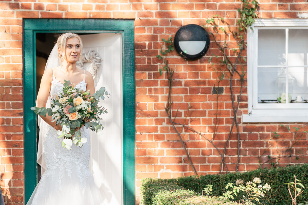 A bride in a white dress and veil stands in a doorway, holding a bouquet of peach and white flowers with greenery. The brick building behind her has a teal door frame and a window with creeping plants. Sunlight creates a warm ambiance.