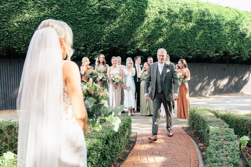 A bride in a white gown and veil approaches a group of smiling people on a garden path. The group includes women with bouquets and a man in a suit, surrounded by greenery and trees.