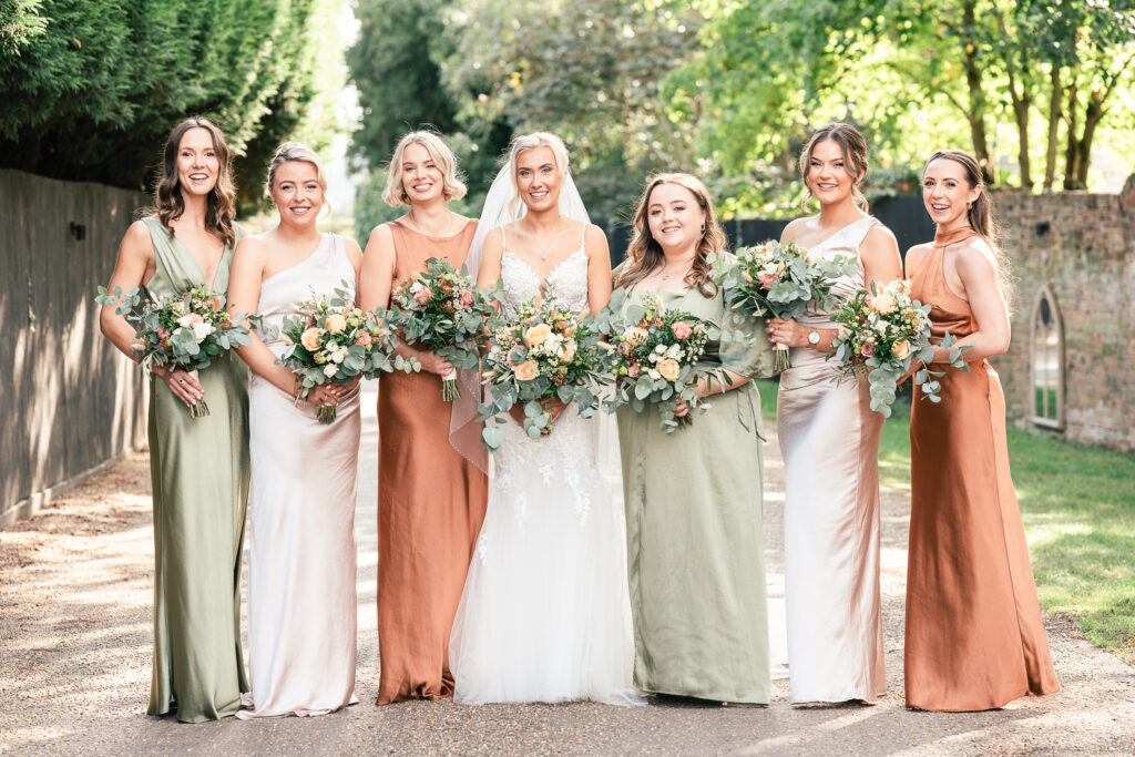 A bride stands with six bridesmaids in a sunlit outdoor setting. The bridesmaids are dressed in earth-toned dresses, varying in shades of green and brown, and hold bouquets of flowers. Trees and a pathway are visible in the background.