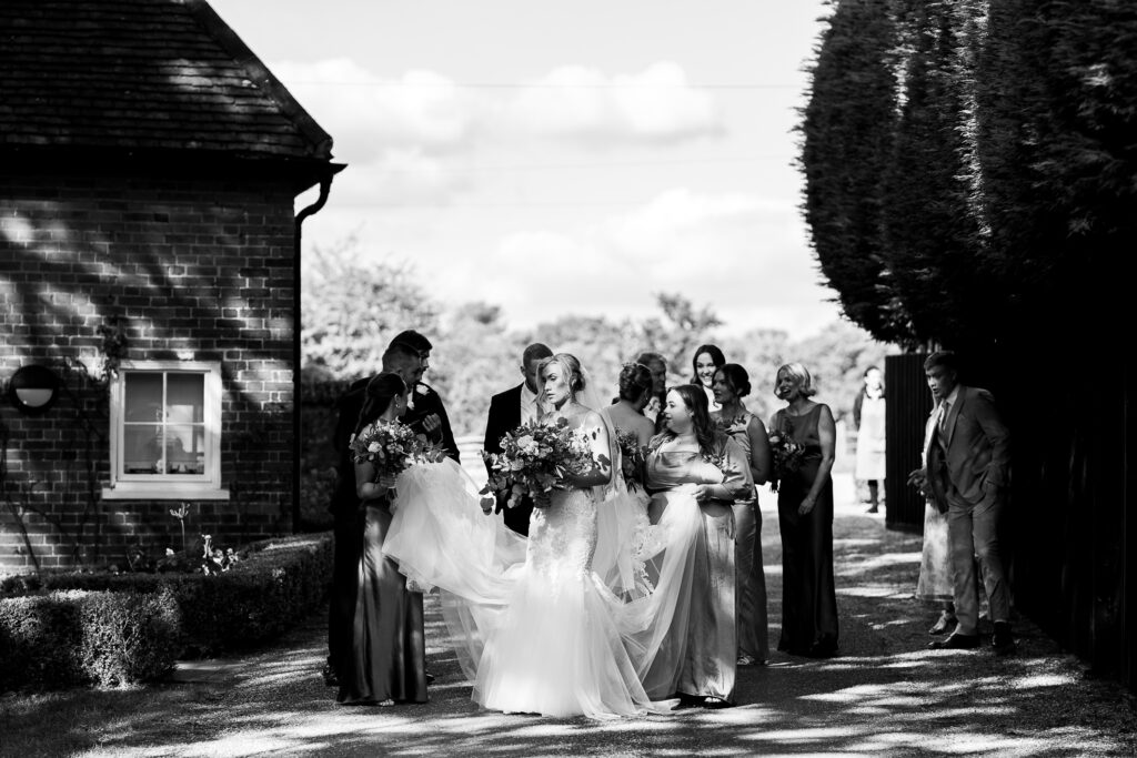 A black and white photo of a bride holding a bouquet, surrounded by bridesmaids in elegant dresses, walking along a garden path near a brick building, with trees casting shadows in the background.