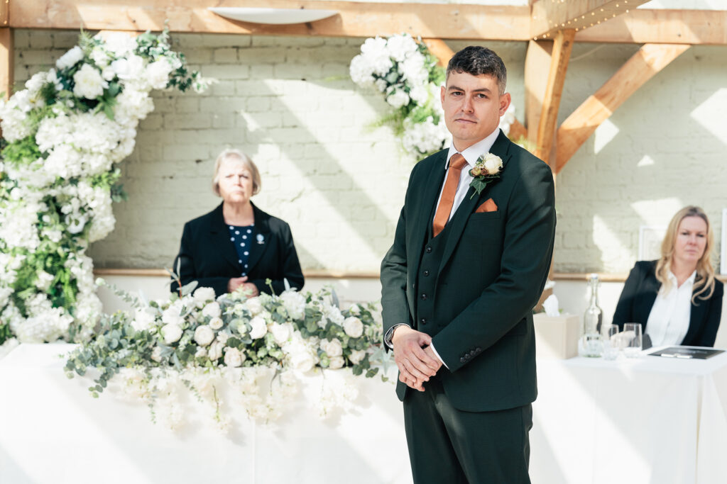 A man in a dark suit stands in front of a flower-adorned table at an indoor event. Behind him are two seated women, one in a dark blazer and another in a light-colored outfit. The room is decorated with white flowers.