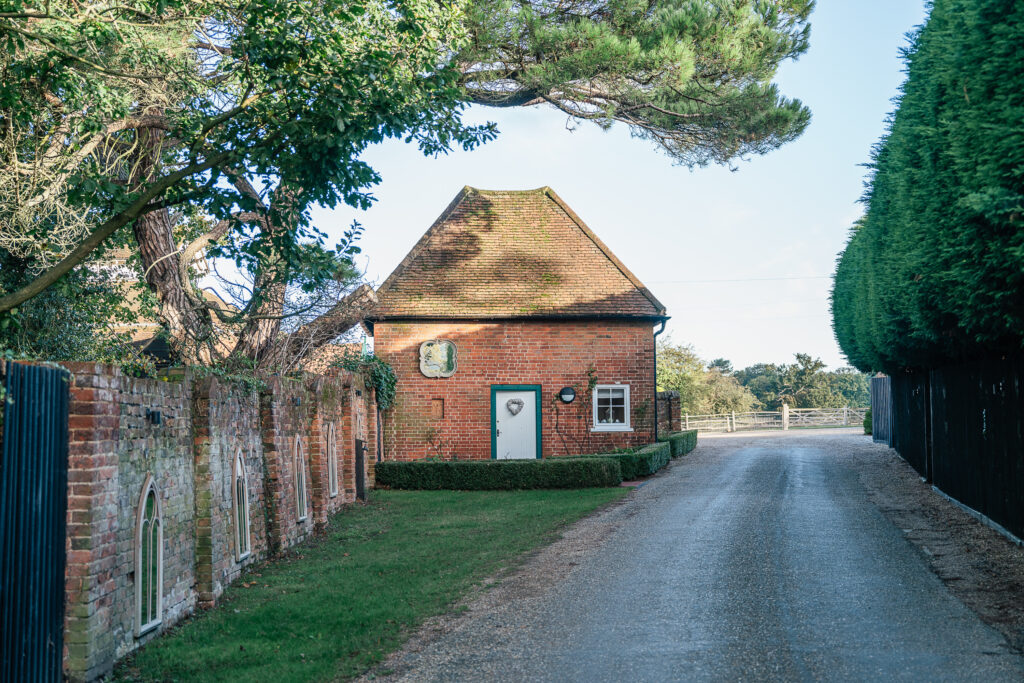 A quaint red brick cottage with a steep roof sits by a quiet road. It's bordered by a neatly trimmed hedge and tall trees, with sunlight filtering through the branches. A low stone wall with arched niches runs along one side.