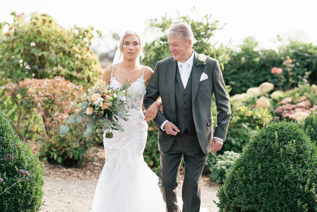 A bride in a white wedding dress holds a bouquet of flowers while walking arm-in-arm with an older man in a suit. They are outdoors, surrounded by lush greenery and bushes.