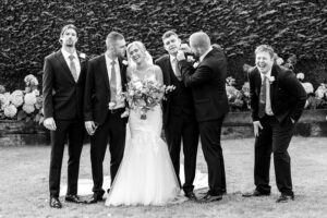 A black and white photo of a bride surrounded by five groomsmen. They are standing on grass with a hedge in the background. The bride holds a bouquet and everyone is smiling or laughing. Gaynes Park, Essex