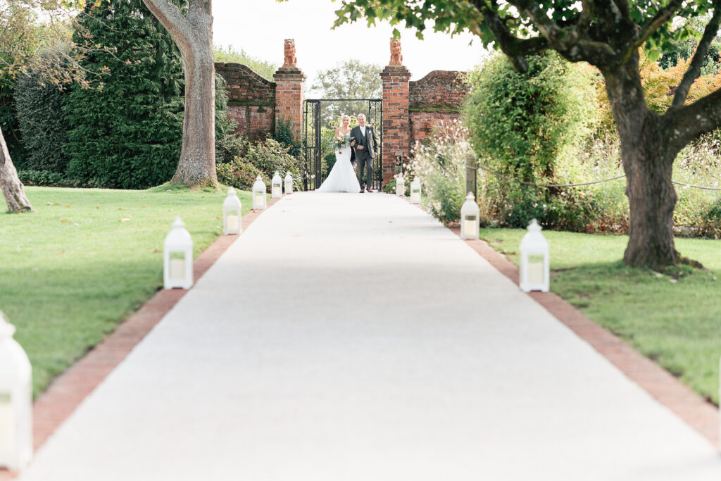 A bride and groom walk hand-in-hand down a long paved path bordered by trees and white lanterns. They are approaching from a decorative brick gate in the background, set in a lush garden scene.