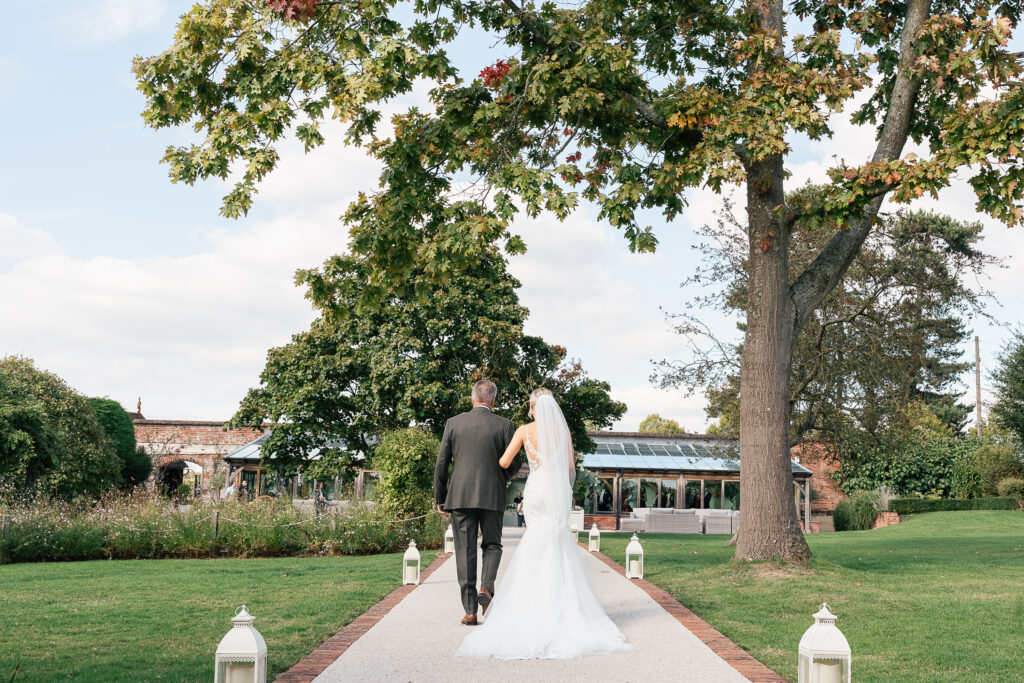 A bride and groom walk hand-in-hand down a garden path under tall trees. The bride wears a white gown and veil, and the groom is in a dark suit. The path is lined with lanterns, and a glass-walled building is in the background.