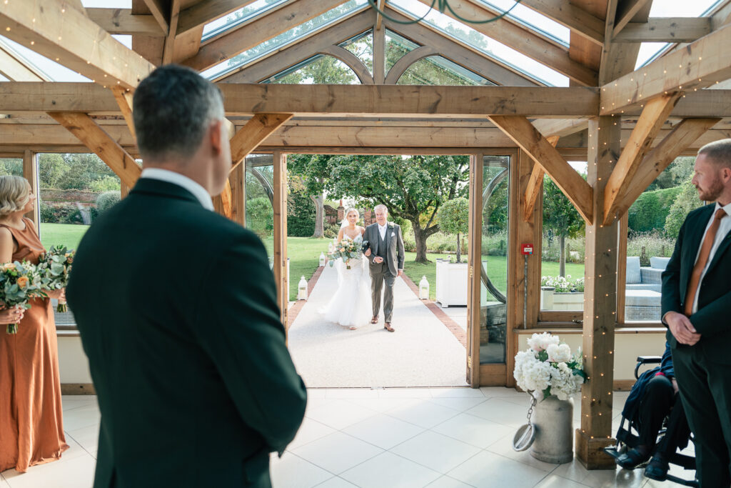 A bride, in a white dress holding a bouquet, walks down the aisle with a man in a gray suit. They're entering a wooden, glass-walled venue. Guests, some holding flowers, watch as the ceremony begins. Greenery and white flowers decorate the space.