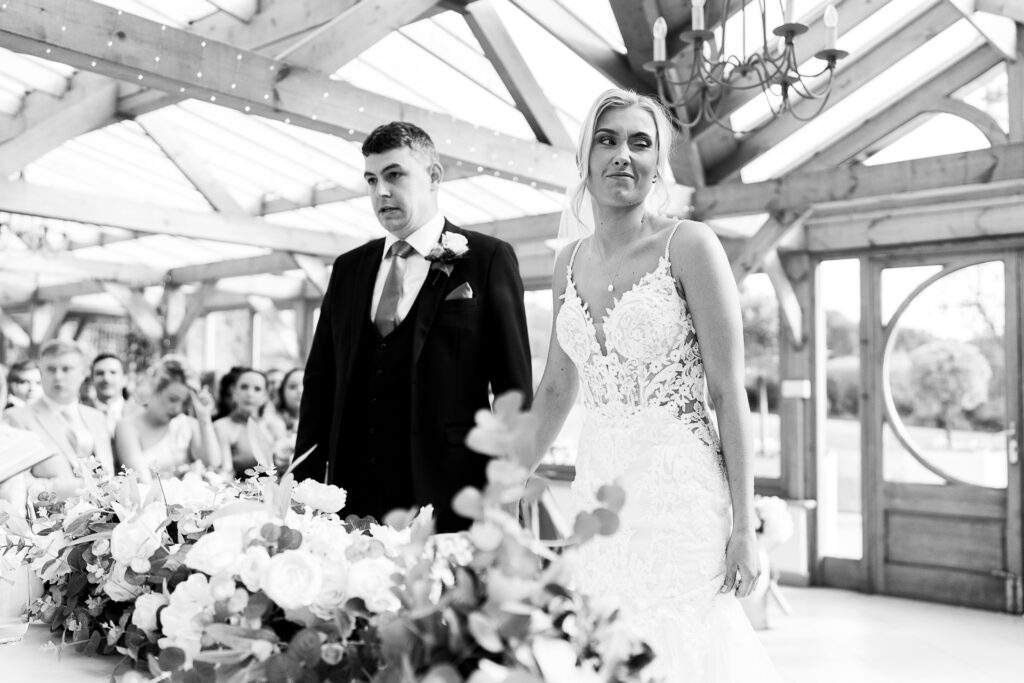 A bride and groom stand together during their wedding ceremony in a bright, wooden venue. The couple is dressed in formal attire, with flowers adorning the table in front of them. Guests are seated in the background. Black and white image.