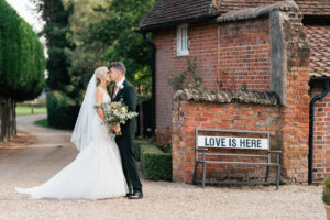 A bride and groom share a kiss outdoors near a rustic brick building. The bride is in a white gown and veil, holding a bouquet, while the groom wears a black suit. A bench next to them displays the sign "LOVE IS HERE." Trees line the pathway at Gaynes Park, Essex