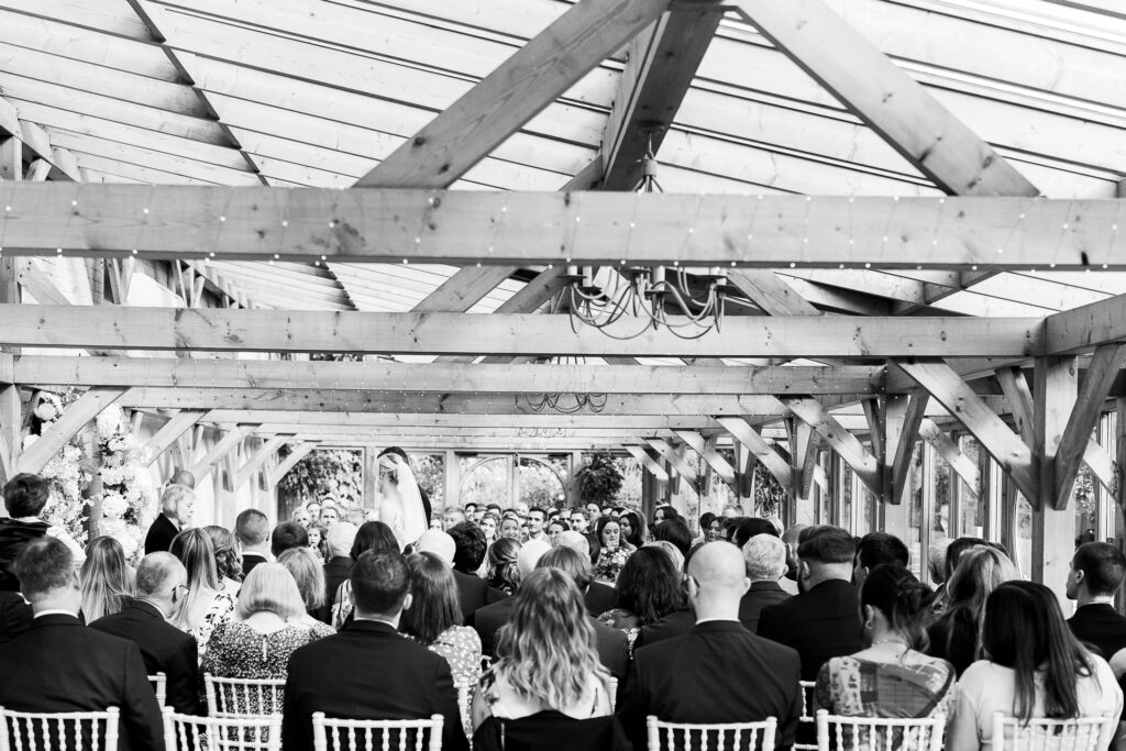 A black and white image of a wedding ceremony in a bright, wooden-beamed venue. Guests are seated on white chairs, facing the couple standing at the altar, with floral arrangements adorning the space.