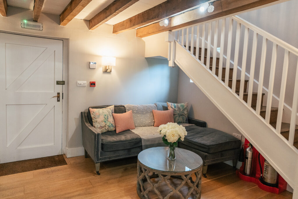 A cozy living room with a gray L-shaped sofa adorned with floral and pink cushions. A round glass coffee table with white flowers sits in front. Wooden beams on the ceiling and a white staircase are visible. Warm lighting creates a welcoming atmosphere.