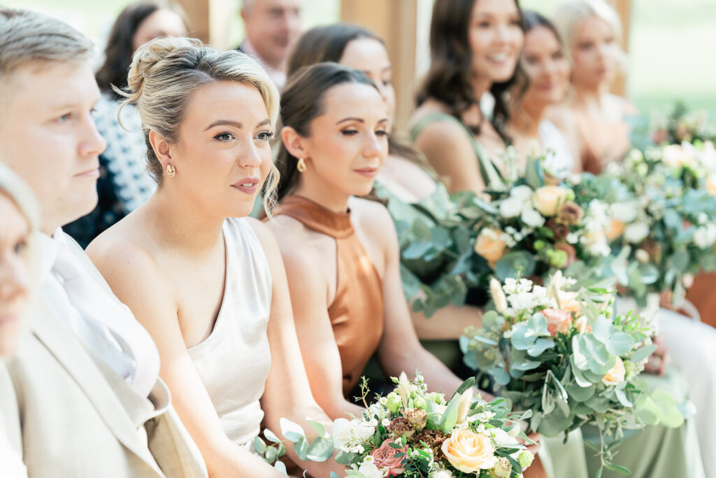 A group of individuals sitting together, wearing formal attire. The women are holding bouquets of flowers with greenery. The scene appears to be at an outdoor event or ceremony.