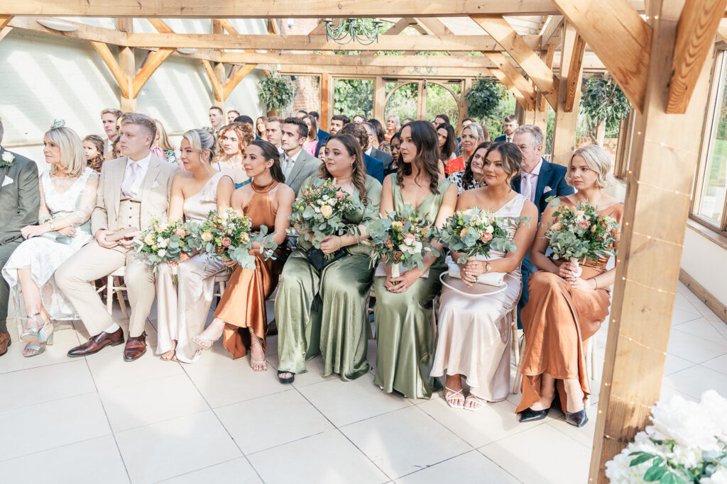 A wedding ceremony with guests seated in a sunlit, wooden-beamed room. Bridesmaids in various shades of green, beige, and copper dresses hold bouquets of green and white flowers. Other attendees sit attentively, dressed in formal attire.