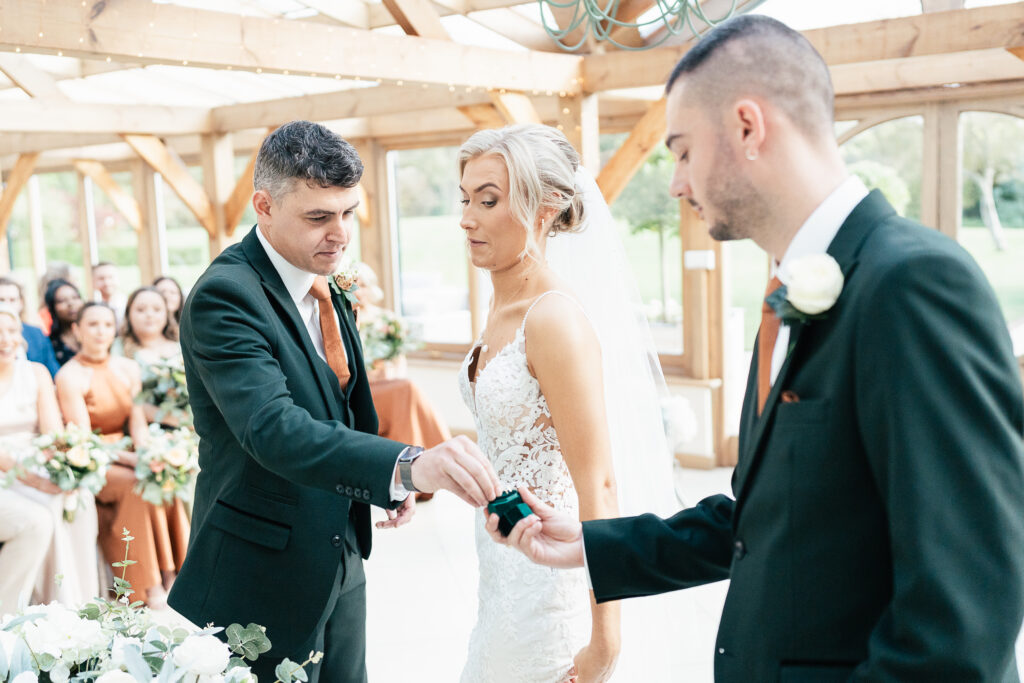 A bride and groom stand inside a bright venue. The groom, wearing a dark suit, is reaching for a ring from a man in a matching suit. The bride wears a white lace dress and veil, looking on attentively. Guests are seated in the background.