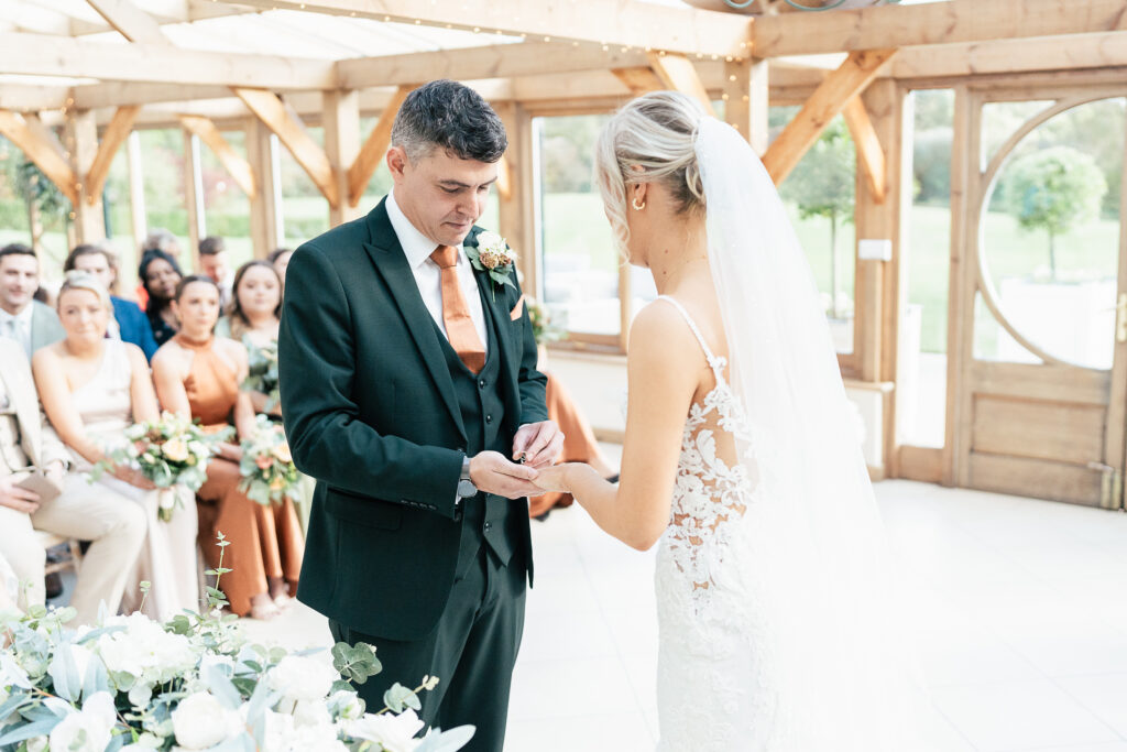 A bride and groom exchange rings during their wedding ceremony in a bright, wooden hall. The groom wears a dark suit with an orange tie, and the bride is in a white gown with a veil. Bridesmaids and guests watch in the background.