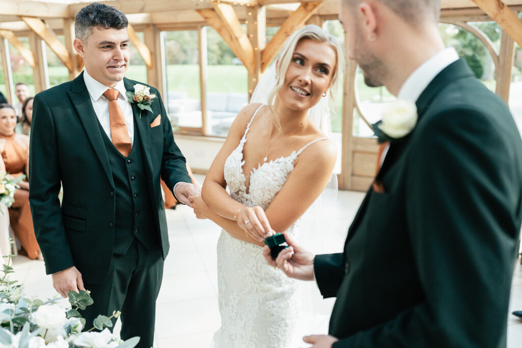 A bride in a white lace dress receives a wedding ring from a man in a black suit. Another man in a black suit stands beside her, smiling. The ceremony takes place in a wooden, sunlit venue with guests seated in the background.