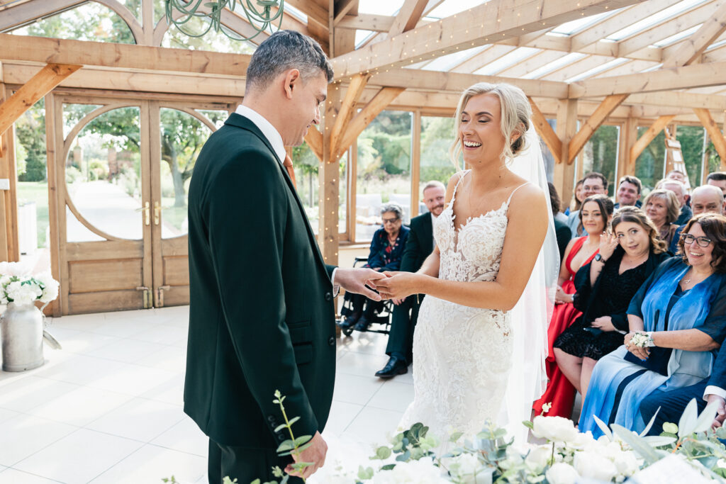 A couple is exchanging rings in a sunlit venue with wooden beams. The bride is wearing a white lace dress and the groom is in a dark suit. Guests, dressed in colorful attire, are seated and smiling in the background.