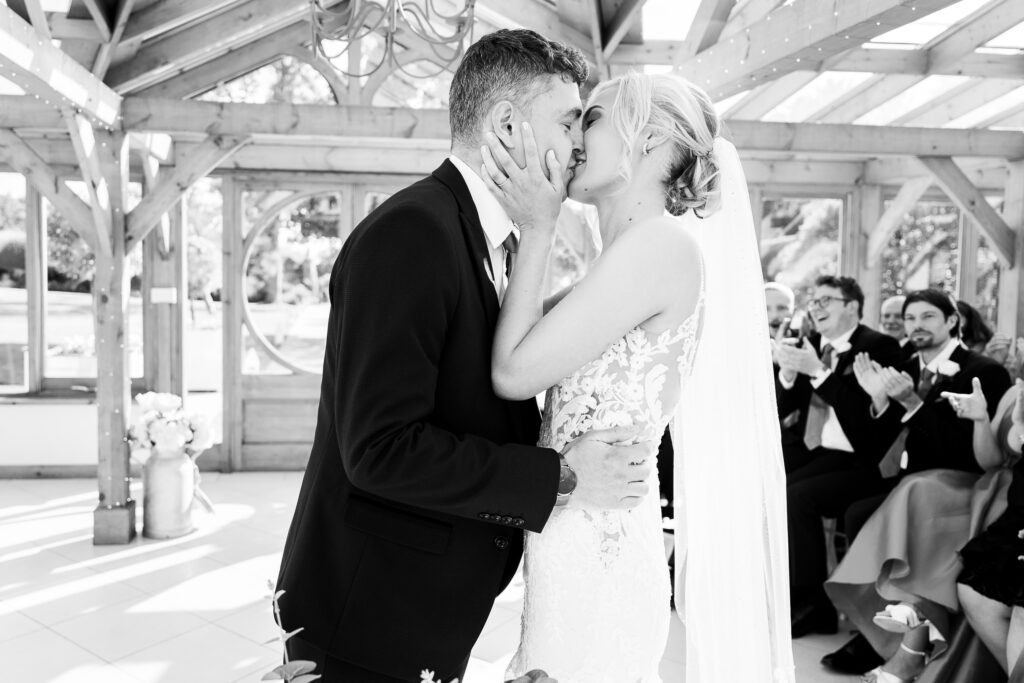 A bride and groom share a kiss during their wedding ceremony in a bright, open venue with wooden beams. Guests in formal attire applaud in the background. The bride wears a lace gown and veil, and the groom is in a suit.