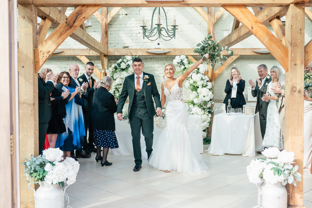 A bride and groom walk down the aisle smiling and holding hands. The bride wears a white gown, and the groom is in a dark suit with an orange tie. Guests around them applaud. The setting features a wooden structure with white floral decorations.