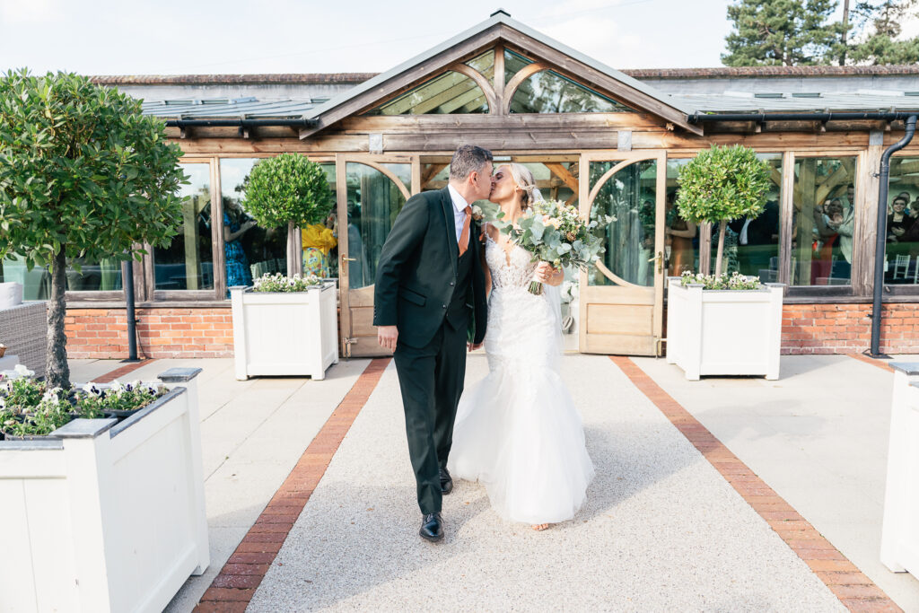 A couple dressed in formal wedding attire walks hand in hand outside a rustic venue. The bride, in a white gown, holds a bouquet, while the groom wears a dark suit. They are surrounded by greenery and white planters on a sunny day.