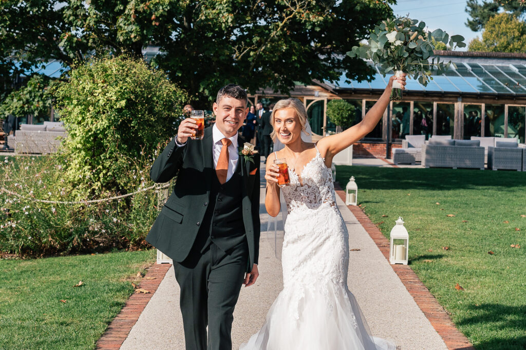 A bride and groom joyfully walk arm in arm down a garden path, each holding a drink. The bride raises her bouquet and smiles. The groom wears a dark suit, and the bride is in a white gown. They are outdoors with greenery and a building in the background.