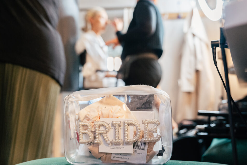 A clear makeup bag labeled "BRIDE" sits in the foreground, filled with items. In the blurred background, a person is having makeup applied by another individual, suggesting a bridal preparation setting.