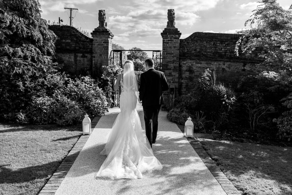 Black and white photo of a bride and groom walking arm in arm down a garden path. The bride wears a flowing gown and veil, and the groom is in a suit. They head toward an ornate wrought-iron gate set in a brick wall, surrounded by lush greenery.