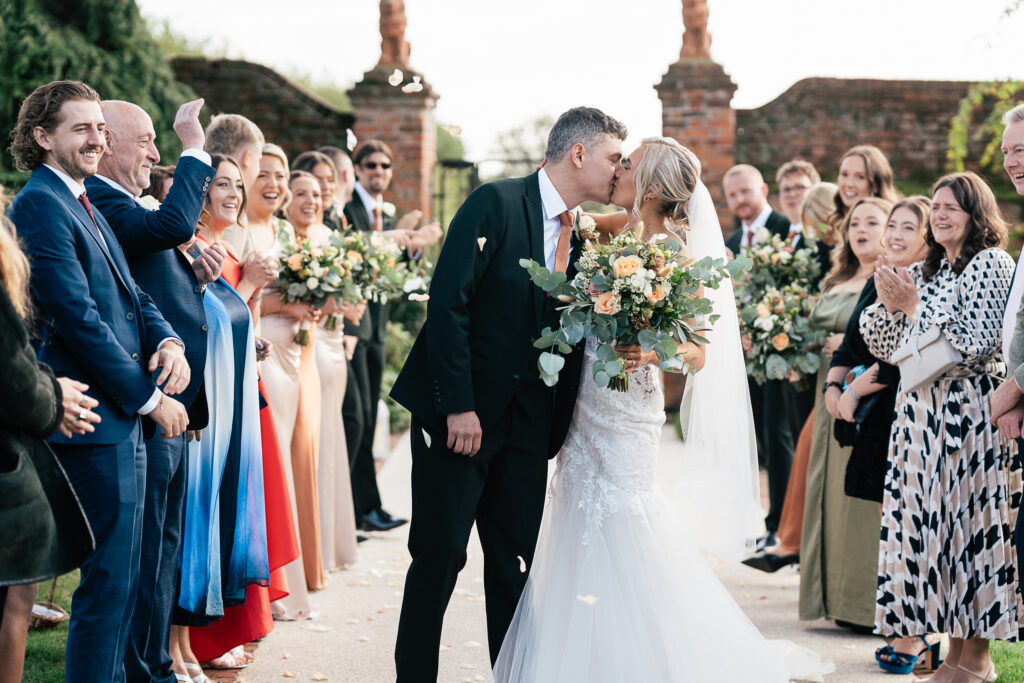 A bride and groom share a kiss amid a cheering crowd of well-dressed guests. The bride holds a bouquet of flowers, and petals are scattered on the ground. The scene is joyful, with people clapping and smiling in celebration.