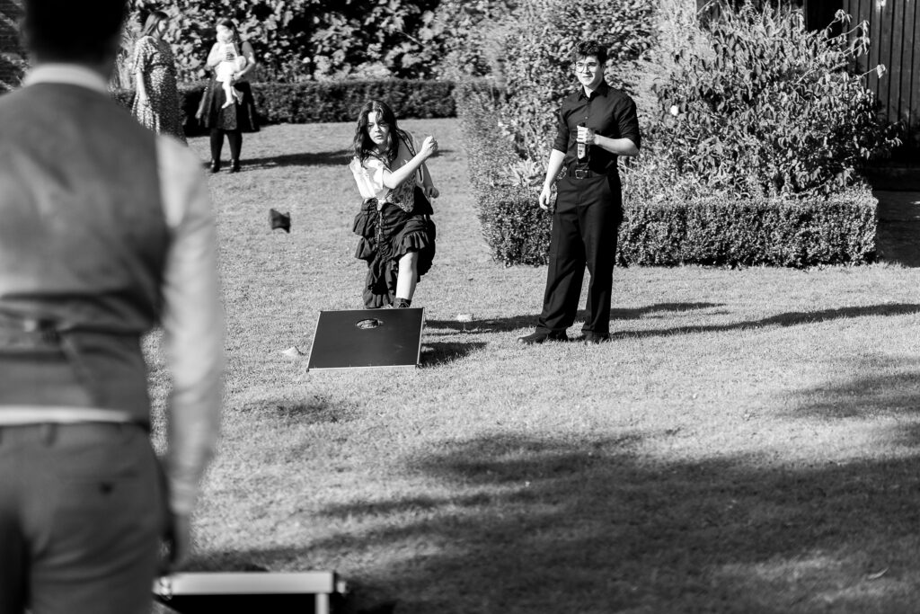 A black and white image of three people in a garden playing cornhole. A woman is throwing a bean bag, while two men watch. Bushes and trees are in the background, and the mood appears light and casual.