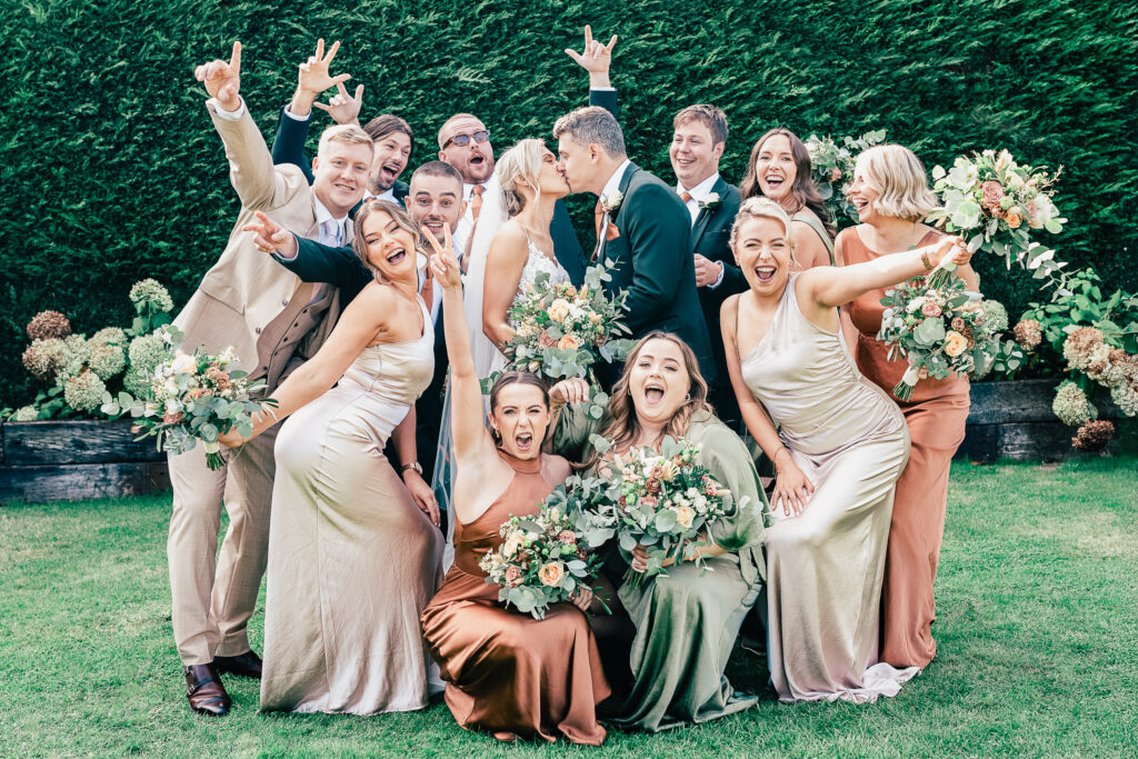 A joyful wedding party poses energetically outdoors. The bride and groom kiss at the center, surrounded by bridesmaids in pastel dresses and groomsmen in beige suits. Everyone smiles and cheers, holding vibrant floral bouquets against a green hedge backdrop.