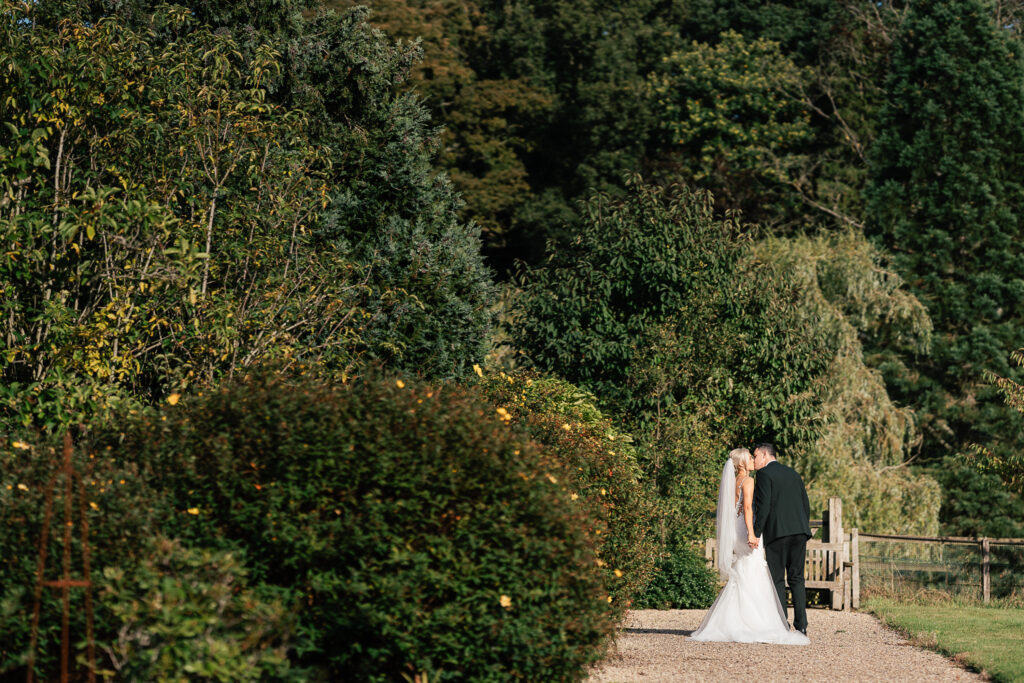 A bride and groom share a kiss on a gravel path surrounded by lush greenery and tall trees. The bride is in a white gown and veil, and the groom is in a dark suit. The scene is serene and romantic.