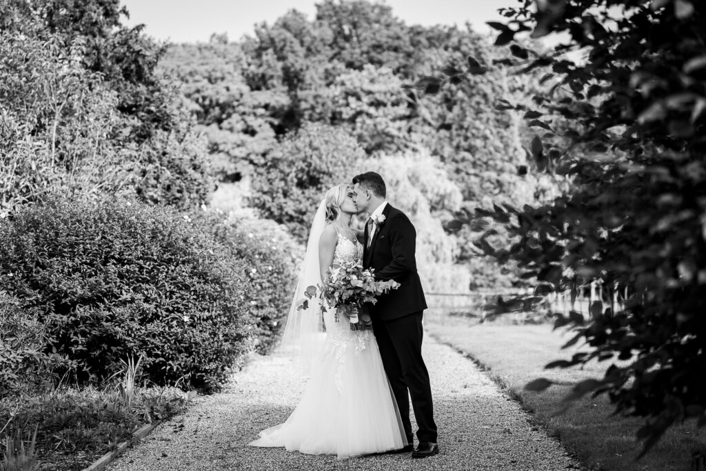 A bride and groom stand on a gravel path surrounded by lush foliage. The bride holds a bouquet, and they share a kiss, creating a romantic moment in a serene garden setting. The image is in black and white.