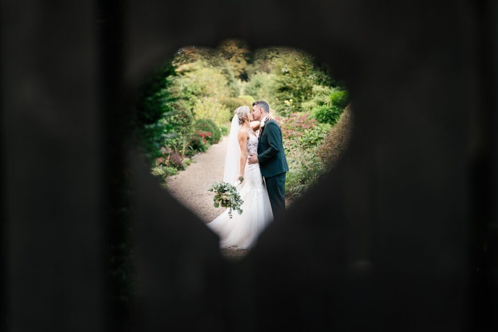 A bride and groom are kissing on a garden path, visible through a heart-shaped opening in a wooden fence. The bride holds a bouquet, and greenery surrounds them.