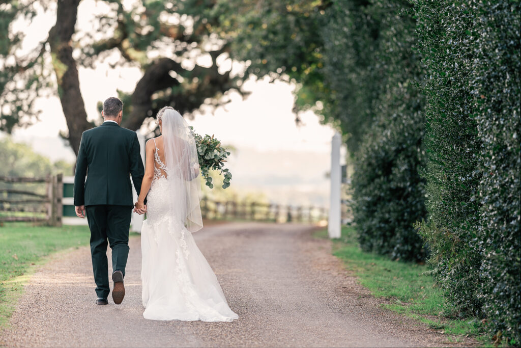 A bride and groom walk hand in hand down a tree-lined path. The bride wears a white gown and veil, while the groom is in a dark suit. Lush greenery surrounds the couple, creating a serene and romantic setting.